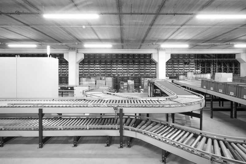 Roller conveyors curving across a warehouse floor, with walls of storage bins and stacked cartons behind them.
