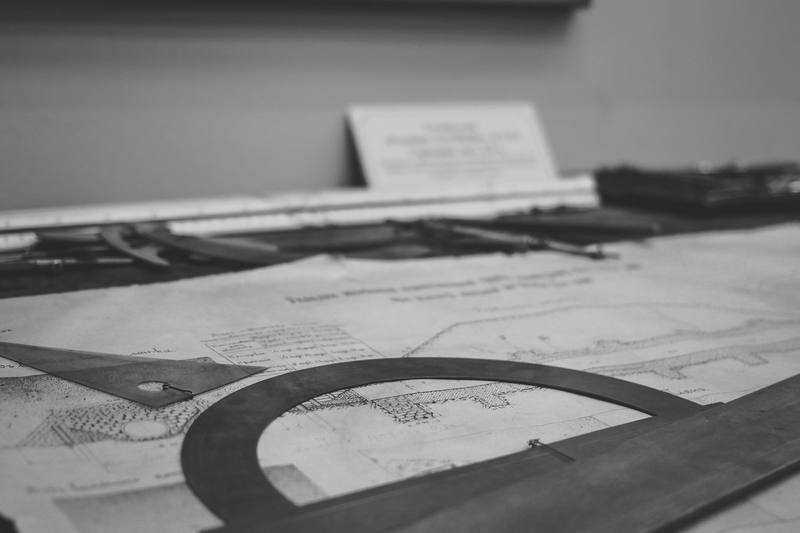 A technical drawing on a desk, with a protractor and a set square lying on it and drafting tools behind.