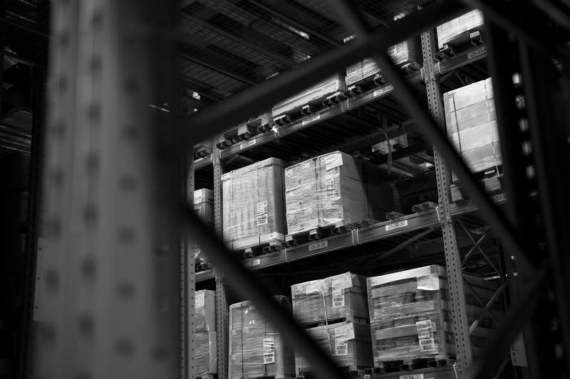 Warehouse pallet racking seen between steel uprights, with stacked cardboard cartons on the shelves.